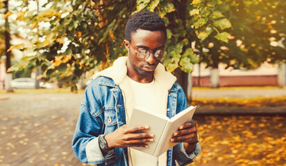 Fototapeta premium Portrait of young african man student reading a book wearing eyeglasses in autumn city park