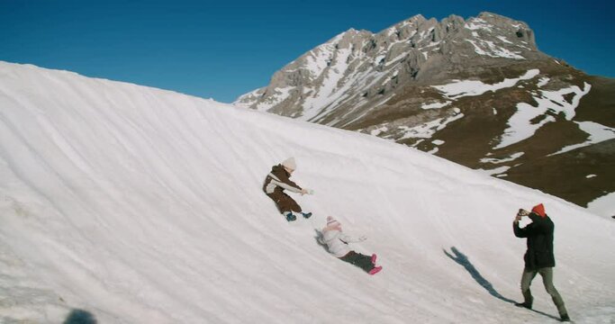 Two children on winter vacation trip roll down a snow slide on their asses with epic mountain landscape and father take photo or video using mobile phone. Family have fun on winter holiday or weekend