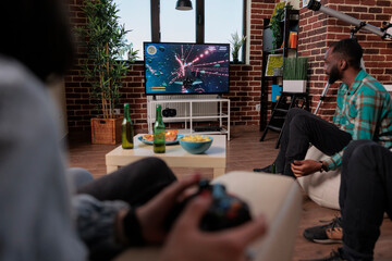 Men and women using tv console and joystick to play video games, having fun at house gathering with...