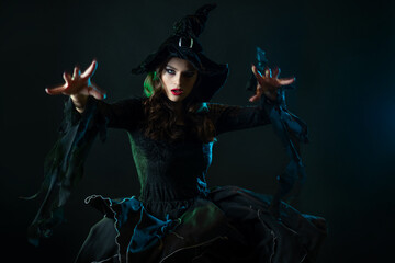 A charming young witch in a costume makes magical passes with two hands, a caster. Young brunette in pointed hat and black dress, photo on black - 532302060