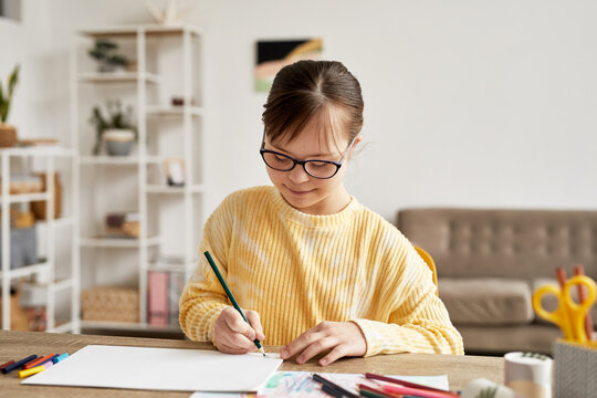 Portrait of teenage girl with Down syndrome drawing pictures at table in cozy room and smiling, copy space