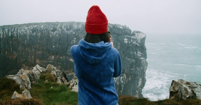 Trendy hipster woman on the cliff ocean shore filming or photographing stormy waves using smartphone on autumn vacation journey. Inspired female travel blogger or influencer make digital content.