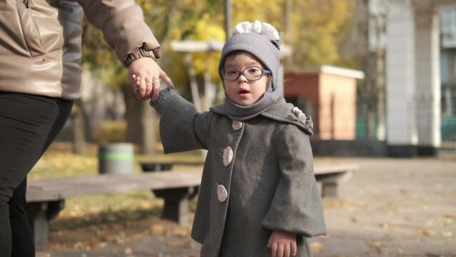 Beautiful little girl in fashionable gray coat and hat and in stylish eyeglasses holding her mommy's hand, family taking a walk along city, attending classes for children with special syndromes