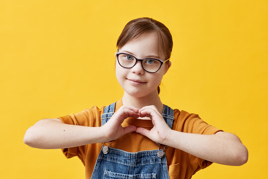 Waist up portrait of teen girl with Down syndrome looking at camera and showing heart sign while standing against yellow background in studio