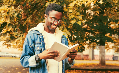 Portrait of happy smiling young african man student reading a book wearing eyeglasses in autumn city park