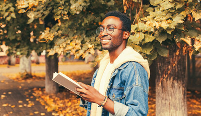 Portrait of happy smiling young african man student reading a book wearing eyeglasses in autumn...