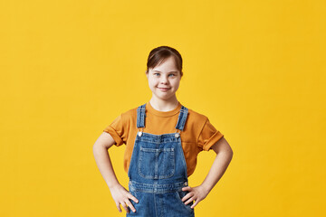 Waist up portrait of cute girl with Down syndrome looking at camera against yellow background in...