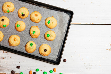 Halloween sugar cookies with chocolate candies in colors orange and green on baking sheet on white background, easy and fun biscuit baking recipe for kids for trick or treating sweets - 532309497