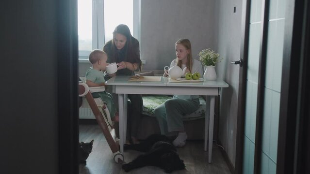 A sweet family of mother and two daughters drink tea in the kitchen in the morning - their pet dog and cat lying on the floor by the table