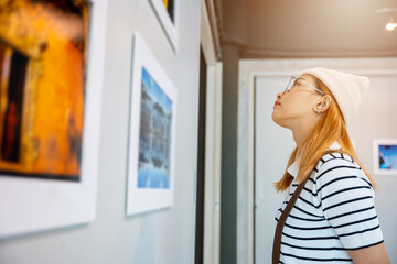 Asian woman standing she looking art gallery in front of colorful framed paintings pictures on...