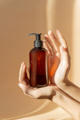 Women's hands hold bottle with dispenser for liquid soap, shampoo, lotion. Mock-up of an unmarked container in sunlight on beige background. Skin care concept, vertical image