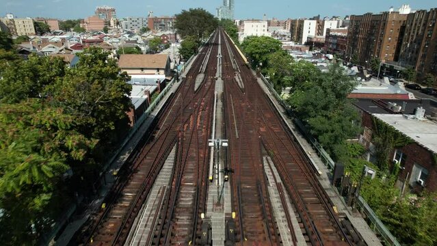 An aerial view of empty, elevated train tracks on a sunny day. The drone camera dolly in over the center of the tracks running through the commercial and residential neighborhood in Brooklyn, NY.