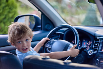 Shaggy child is holding steering wheel in his father's car like real driver. Boy amusingly plays driver of a real car. Five-year-old boy gets behind the wheel and pretends to drive a car