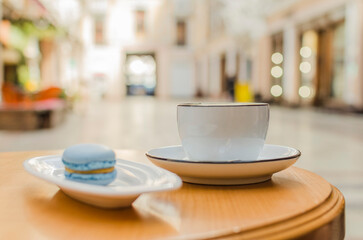 A white cup with a black border stands on a saucer, next to it is a blue macaroon on a plate