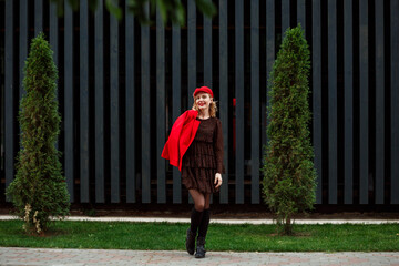 Full length portrait of fashionable woman in red cap, red jacket, dark dress, black boots with gaiters in front of modern wooden building outdoors in the city streets. 