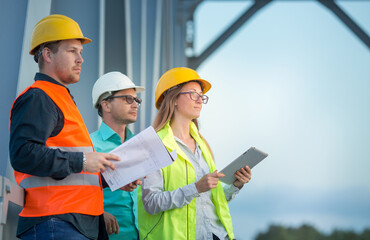 Railway and road engineers are standing in orange and white helmets and signal vests with documents against the background of the metal structure of the bridge.