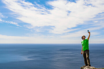 Man standing on an edge of a rock and taking pictures on smart phone of vast ocean and blue cloudy...