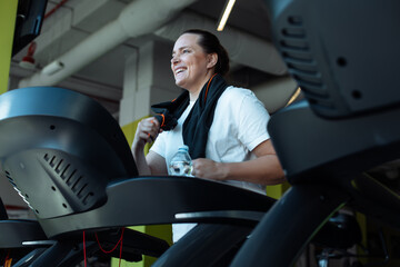 Smiling overweight woman vigorously run on treadmill at fitness gym with towel hanging around neck,...