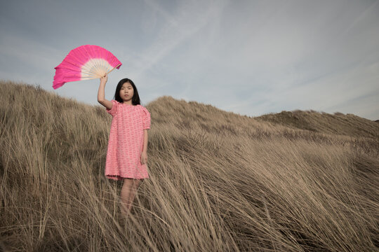 portrait of asian girl in pink dress standing in landscape of  dunes holding chinese fan