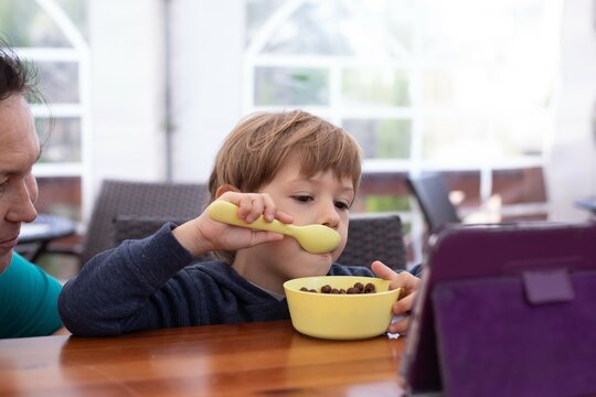 Little boy sit on chair at table and watch cartoon on tablet computer with mother. Kid of kindergarten age eat chocolate balls with milk from children tableware. Ready breakfast, gadget addiction
