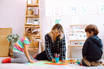 Kind teacher conduct lesson for little children while sitting on floor in playing room. Kids play with toys and draw picture with colored pencils in kindergarten. Lesson, preschool, daycare