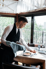 A young male waiter in a stylish uniform is engaged in serving the table in a beautiful gourmet...