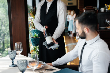 A young sommelier in a stylish uniform demonstrates and offers the client fine wine in the restaurant. Customer service. Table setting in a fine restaurant.