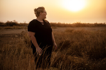Beautiful overweight woman of xl size dressed in a black dress posing in a field with dry grass against the background of a magnifivent sunset