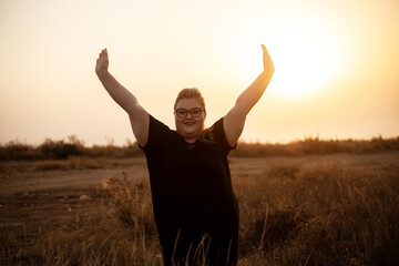 Beautiful overweight woman of xl size dressed in a black dress posing with her hands raised in a field with dry grass against the background of a magnifivent sunset