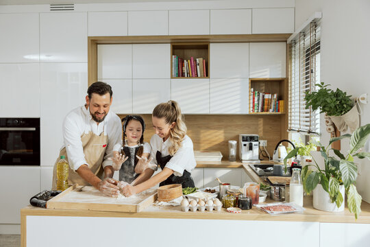 Happy family kneading dough together teaching daughter cooking baking homemade pizza. Pizza ingredients on wooden surface eggs flour tomato sauce olives asparagus salami mushrooms oil.