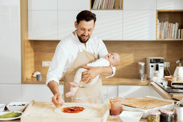 Fototapeta na wymiar Adorable young father with lovely son in hands preparing homemade pizza making surprise for wife applying tomato sauce on homemade dough standing in modern kitchen.