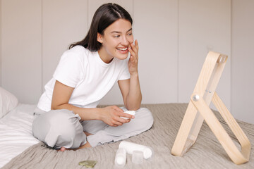 Positive young caucasian girl applying moisturizing cream on face and smiling with teeth in morning. Brunette hair wear pajamas sitting on bed at home. Concept beauty lady, cosmetic.