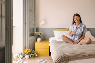 Happy young caucasian woman with wide smile looks at camera and sits on bed near window. Girl with brunette hair holds glass of water in her hand. Happy morning concept.