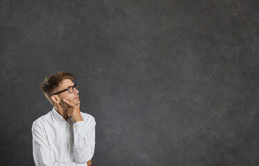 Young dreamy male thinker pensively looks at free space for text on gray background. Serious Caucasian man in white shirt and glasses holding his chin ponders something important. Advertising banner.