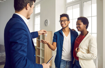 Obraz premium Buying home. During meeting with happy young married couple realtor gives them keys to their new home. Real estate agent in formal suit has signed successful contract with African-American couple.