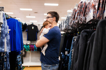 Young dad in shopping centre looking at winter clothing holding a baby