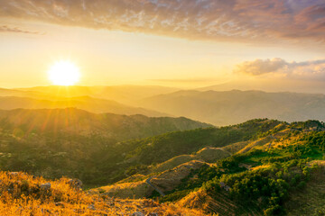 Mountain valley during sunset or sunrise. Rocks and slopes covered with green trees with amazing cloudy sunny sky on background.