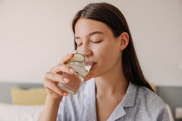Beautiful young caucasian girl closes her eyes and drinks water with slice of cucumber morning. Woman with brown hair in hotel room. Concept of beauty, lifestyle.