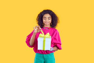 Little child gets a cool birthday present. Happy kid opening a beautiful gift box. Pretty Afro American girl standing isolated on a yellow background, holding a white box and untying the bow