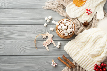 Christmas Eve concept. Top view photo of hat mittens scarf cup of cocoa with marshmallow placemat reindeer ornament anise cinnamon sticks dried orange slices viburnum on grey wooden desk background