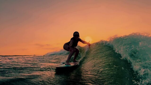 Silhouette of surfing sporty girl surfing with shaka on wave on lake on the setting orange sun