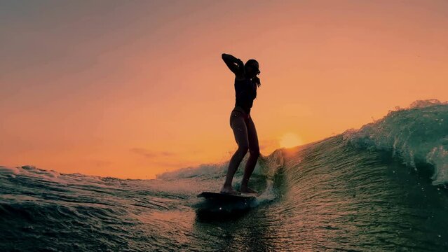 Silhouette of a dancing sporty girl surfing on wave surface on the lake on the setting orange sun
