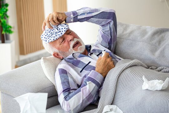 Mature man feeling sick with cold and fever at home, ill with flu disease sitting on the sofa with ice pack on his head