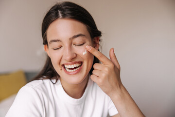 Close-up happy young caucasian woman using face cream and laughs with closed eyes. Brunette hair girl in white t-shirt at home. Concept positive emotions, skin care.