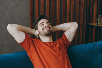 Young man wear red t-shirt listen music in earphones hold hands behind neck sit on blue sofa couch...