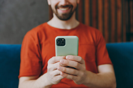 Cropped young man wear red t-shirt hold in hand use mobile cell phone sit on blue sofa couch stay at home hotel flat rest relax spend free spare time in living room indoors grey wall Focus on device