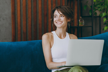 Young IT woman wear white tank shirt look aside hold use work on laptop pc computer sit on blue...