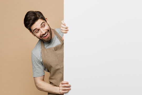 Young man barista barman employee in brown apron work in coffee shop look at big white billboard for content place text image isolated on plain pastel beige background Small business startup concept