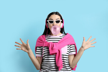 Fashionable young woman with headphones blowing bubblegum on light blue background