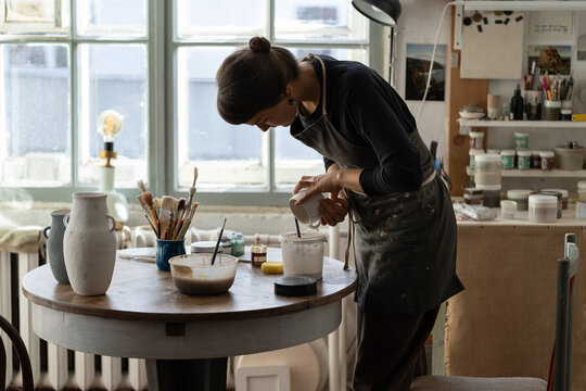 Young female artisan stands near round wooden table with equipment for work with pottery against bright big window. Brunette woman in black apron pours white paint into ceramic cup in workshop